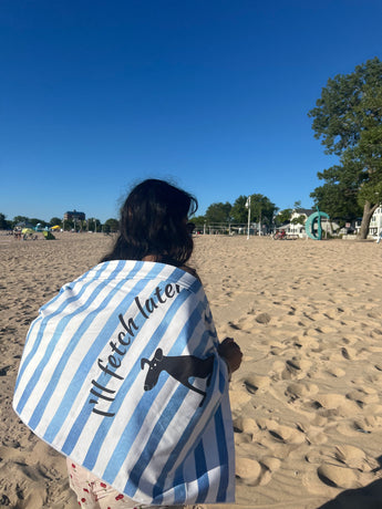 Person on a sandy beach wrapped in a blue and white striped Pouty Paws dog towel with a black dog silhouette